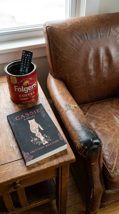 Book titled 'Cassie' by Brittany Williams on a wooden table next to a can of Folgers coffee and a remote control, with a brown leather couch in the background.